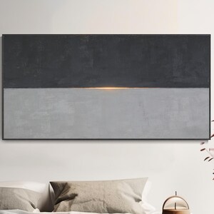 Puede incluir: Arte mural abstracto con un lienzo rectangular dividido horizontalmente. La mitad superior es gris oscuro, la inferior gris claro, con una fina línea dorada. La obra de arte se exhibe sobre una cama.