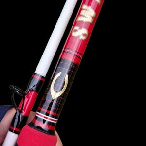 May include: Red and black fishing rod with gold lettering that reads "SWAMS". The rod has a white pole and a red and black handle. The rod is held against a black background.