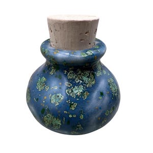 Può includere: Un piccolo vasetto in ceramica rotondo con una smaltatura blu lucida caratterizzata da macchie verdi e gialle. Il vasetto presenta un bordo largo e arrotondato ed è sigillato da un tappo di sughero cilindrico color marrone chiaro.