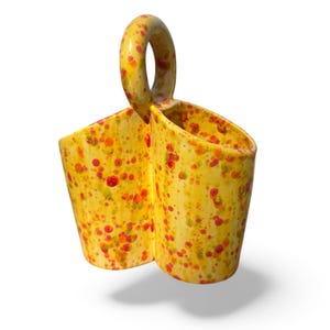 May include: A yellow ceramic utensil holder with a handle, featuring a speckled pattern of red, orange, and green dots. The holder has two compartments and is designed for kitchen use.