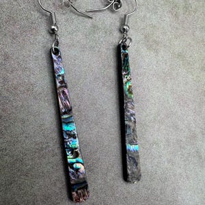 May include: A pair of dangle earrings with iridescent abalone shell inlays. The earrings have a rectangular shape with a silver-tone hook. The abalone shell displays shades of blue, green, purple, and brown.
