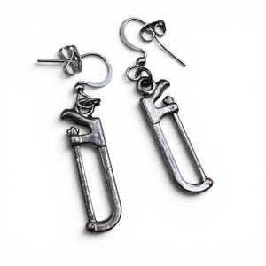 May include: A pair of silver-colored earrings shaped like miniature hand saws. The earrings have a detailed design, including the saw blade and handle. The hooks and posts are also silver. The earrings are displayed against a white background.