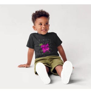 Puede incluir: Una camiseta negra con el texto "Never Playing" en rosa y blanco, con un diseño de corona verde. El niño lleva pantalones cortos verde oliva y zapatillas blancas.