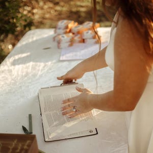 May include: A person is holding open a book with text on a white table. The book is black with a silver clasp. There are small, wrapped packages in the background. The person is wearing a white dress.