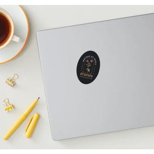 May include: A sticker on a laptop with the text "INSPIRED BY GRACE STUDIOS" and a floral design. A cup of coffee, a yellow pen, and two gold paper clips are also on the white surface.