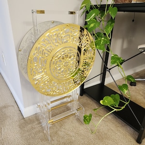 May include: A gold decorative stand with a round top and three legs. The stand is made of clear acrylic and has a gold metal top. The top is decorated with a circular design of intricate patterns.