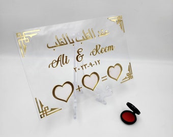 May include: A clear acrylic sign with gold lettering and decorative borders. The sign reads "Ali & Reem" with the date "2023-9-12" and two hearts with a plus sign and an equals sign. The sign is on a white background.
