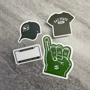 May include: A collection of Sacramento State Hornets fan merchandise, including a baseball cap, a t-shirt sticker with "SAC STATE 1947", a license plate frame, and a foam hand with "Stingers Up!" text. The items are in green and white.