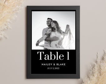 Puede incluir: Un cartel de mesa de boda en blanco y negro con el texto "Table 1" en una fuente blanca grande. Debajo del número de mesa, los nombres "Hailey & Blake" y la fecha "July 5, 2022" están impresos en una fuente blanca más pequeña. Una foto en blanco y negro de una pareja está en el fondo.