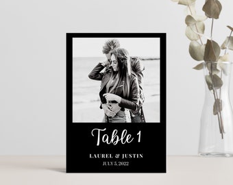 Puede incluir: Tarjeta de número de mesa de boda en blanco y negro con una foto de una pareja en el fondo. La tarjeta dice "Mesa 1" en una fuente blanca cursiva y "Laurel & Justin" y "5 de julio de 2022" en una fuente blanca más pequeña.