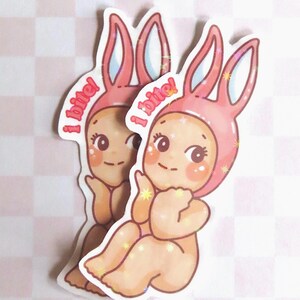 May include: Two stickers featuring a cartoon baby wearing a pink bunny hat. The baby has rosy cheeks and is surrounded by glitter. The text "I bite!" is written above the baby's head in pink letters.