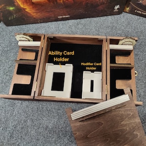 Puede incluir: Una caja de madera con compartimentos para guardar cartas de juego. La caja tiene dos secciones, una etiquetada como "Porta tarjetas de habilidad" y la otra como "Porta tarjetas de modificador".