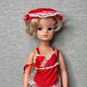 May include: A vintage doll with blonde hair wearing a red and white lace dress and a red hat with white lace trim.