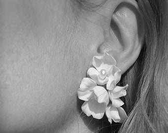 以下が含まれることがあります： 複数の花びらが重なり合って房状になった、花の形のスタッドピアスのモノクロ写真。一番上の花の中央には、小さく丸い滑らかなビーズがあしらわれています。ピアスは耳たぶに装着されています。