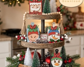 Könnte beinhalten: Ein dreistöckiges Holztablett, weihnachtlich dekoriert mit Miniatur-Holzschildern, die einen Weihnachtsmann, Elfen, Schneemann und Pinguin zeigen. Das Arrangement enthält kleine Flaschenbürstenbäume, Tannenzapfen, rote Kugeln und Beeren.