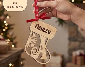May include: A light-colored wooden Christmas stocking ornament featuring a cutout snowflake design and the name Ashley engraved on the cuff. The ornament is suspended by a red ribbon with white snowflake patterns. Text reads: BUNDLE of 26 DESIGNS.