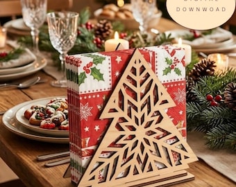 May include: A light-colored wooden napkin holder featuring a laser-cut Christmas tree design with a geometric snowflake pattern. The holder contains a stack of red paper napkins decorated with green holly leaves and white star motifs.