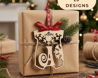 May include: A light-colored wooden ornament featuring a cutout snowflake design and the name Alex above a large letter A. The ornament is tied with a red ribbon and hangs on a brown gift box. A text bubble in the corner reads BUNDLE of 26 DESIGNS.