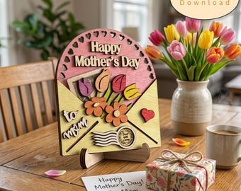 May include: A wooden sign shaped like an envelope with the text Happy Mother's Day and to mom, featuring cutouts of flowers and a heart. The sign includes a stamp graphic and sits on a wooden stand, accompanied by a gift box and a card.