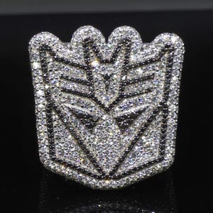 Könnte beinhalten: Ein silberfarbener Ring mit einem schildförmigen Emblem, das mit zahlreichen kleinen, klaren, funkelnden Steinen besetzt ist. Das Design zeigt ein stilisiertes Robotergesicht mit schwarzen Akzenten, die die geometrischen Formen betonen.