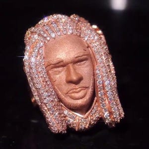 Puede incluir: Un anillo de oro rosa que presenta un rostro humano esculpido con cabello trenzado. El cabello está incrustado con numerosas piedras pequeñas, claras y brillantes que captan la luz. Los rasgos faciales tienen un acabado mate que contrasta.