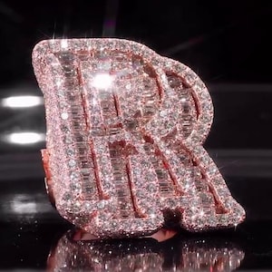 May include: A rose gold ring in the shape of the letter "R", encrusted with numerous small, sparkling gemstones. The ring is set against a dark background, with light reflecting off the gemstones, creating a dazzling effect. The ring is a statement piece.