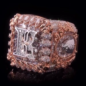 Puede incluir: Un anillo estilo campeonato en oro rosa con una gran letra K central incrustada con pequeñas piedras blancas. El anillo está decorado con intrincados detalles metálicos, piedras blancas adicionales y un emblema lateral circular con un signo de dólar.