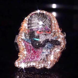 Puede incluir: Un anillo grande con una figura central adornada con numerosas piedras preciosas pequeñas y brillantes en blanco, rosa y azul. La figura lleva un tocado detallado y está rodeada por un borde denso de piedras engastadas en oro rosa.