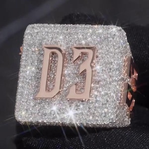 Puede incluir: Un anillo cuadrado con una superficie brillante similar a un diamante. El anillo presenta las letras "D3" en oro rosa. El anillo se muestra sobre un fondo oscuro, resaltando su brillo.