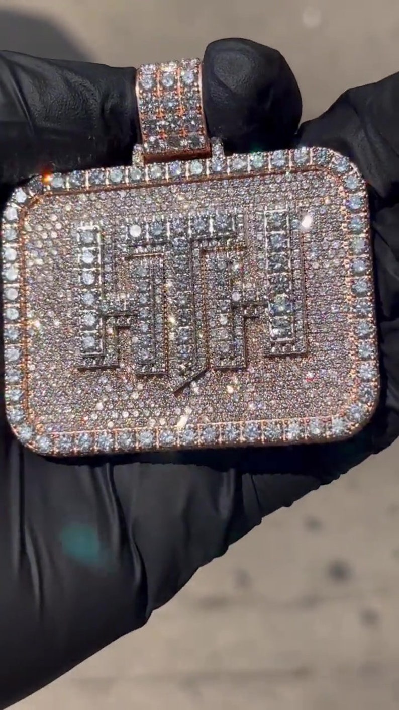 K&ouml;nnte beinhalten: Ein goldener Anh&auml;nger mit quadratischer Form, bedeckt mit kleinen Diamanten, die das Wort "HUF" in fetter Schrift buchstabieren.