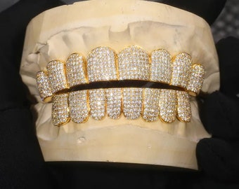 May include: Gold-colored, diamond-studded grillz, displayed on a dental mold. The grillz are designed to cover the upper and lower teeth. The diamonds are small and closely set, creating a sparkling effect. The dental mold is a light beige color.