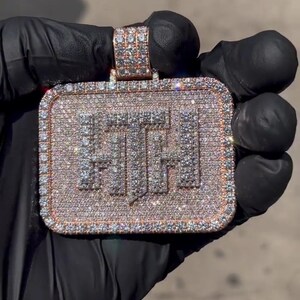 K&ouml;nnte beinhalten: Ein goldener Anh&auml;nger in rechteckiger Form mit den Buchstaben "F.T.H." besetzt mit Diamanten. Der Anh&auml;nger wird in einer schwarzen behandschuhten Hand gehalten.