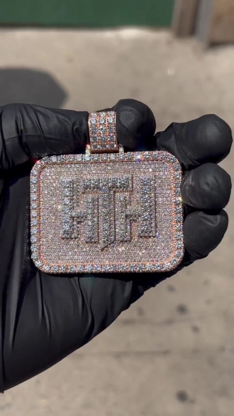 K&ouml;nnte beinhalten: Ein goldener Anh&auml;nger in rechteckiger Form mit den Buchstaben "F.T.H." besetzt mit Diamanten. Der Anh&auml;nger wird in einer schwarzen behandschuhten Hand gehalten.