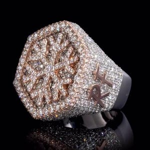 May include: A large, ornate ring with a geometric design and sparkling stones. The ring features a rose gold and silver color scheme, with the initials "R" and "X" on the side. The ring is set against a black background.