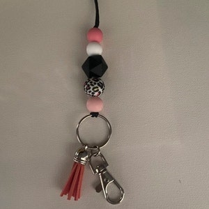 Puede incluir: Un llavero de cuentas de color rosa, blanco, negro y estampado de leopardo con un anillo de llave plateado y un borla rosa.
