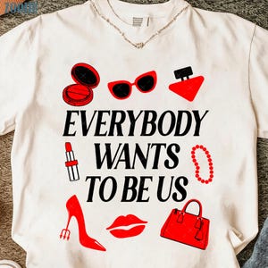 Puede incluir: Camiseta blanca con un diseño gráfico que presenta artículos de moda y cosméticos rojos. El texto "EVERYBODY WANTS TO BE US" está impreso en negro. El diseño incluye lápiz labial, gafas de sol, un bolso y un tacón alto.