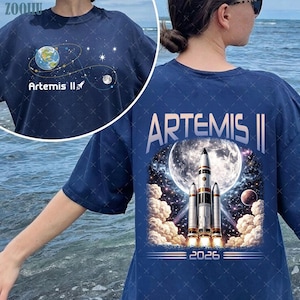 Puede incluir: Camiseta azul marino con diseño espacial. La parte delantera muestra un planeta y estrellas con el texto "Artemis II". La parte trasera muestra un cohete lanzándose hacia la luna, con "Artemis II" y "2026".