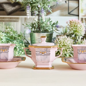 Può includere: Un set di tre tazzine da tè e piattini rosa e dorati con un designo marmoreo. Le tazzine e i piattini hanno un motivo geometrico in oro e nero. Il set include una zuccheriera con coperchio.