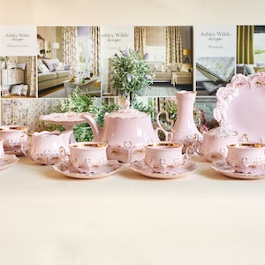 Op de afbeelding: Een roze en gouden theeservies met een theepot, roomkan, suikerpot en vier kopjes en schoteltjes. De set is versierd met delicate bloemmotieven en gouden accenten.