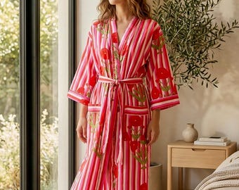 May include: A red and white striped robe with a floral pattern. The robe has a matching belt and pockets. The sleeves are three-quarter length. The robe is knee-length and is worn by a person standing near a window.