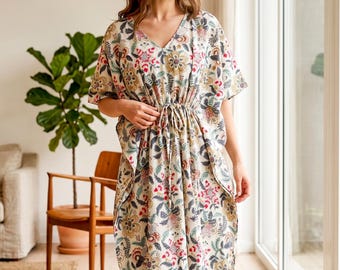May include: A long, flowing, V-neck dress with a floral pattern in red, yellow, and blue on a cream background. The dress has short sleeves and a drawstring waist. The model is standing barefoot near a large window.