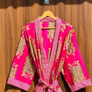 May include: A vibrant pink robe with a tiger print and a striped trim. The robe is made of a lightweight fabric and has a matching belt. The tiger print features orange and black stripes on a pink background. The robe is hanging on a wooden hanger.
