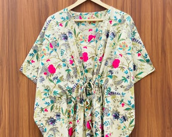 May include: A cream-colored, floral-print kaftan dress with a V-neck and drawstring waist. The dress features a vibrant pattern of pink, blue, and green flowers and leaves. It hangs on a wooden hanger against a wood-grain background.