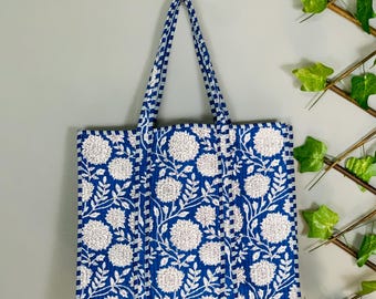 May include: A blue tote bag with a white floral pattern hangs against a gray wall. The bag features a repeating design of white flowers and leaves on a blue background. The handles are blue and white striped.