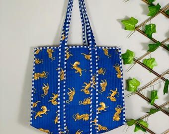 May include: A blue tote bag with a repeating pattern of yellow tigers. The bag has white and blue striped handles and trim. The bag is hanging on a wooden peg against a gray wall.