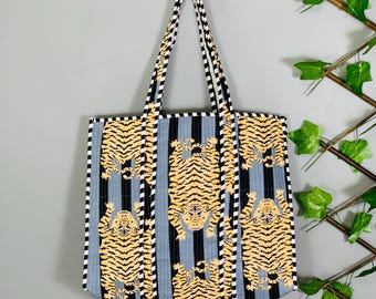 May include: A quilted tote bag with a tiger print. The bag features a pattern of yellow tigers on a blue and black striped background. The handles are black and white striped. The bag is hanging on a wooden hook.