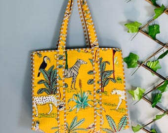 May include: Yellow tote bag with a jungle animal print featuring a zebra, leopard, and toucan. The bag has matching handles and trim. The bag is hanging on a gray wall.