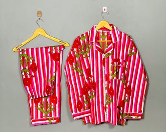 May include: A two-piece pajama set with a red and white striped pattern. The set includes a button-down shirt and matching pants, both adorned with a floral print in red and green. The pajamas are hanging on wooden hangers.