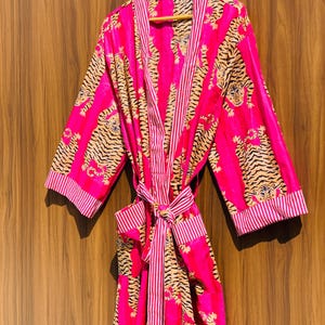 May include: A vibrant pink robe with a tiger print design. The robe features a striped trim and a matching belt. The tiger pattern is in shades of tan and black. The robe is hanging on a wooden hanger.