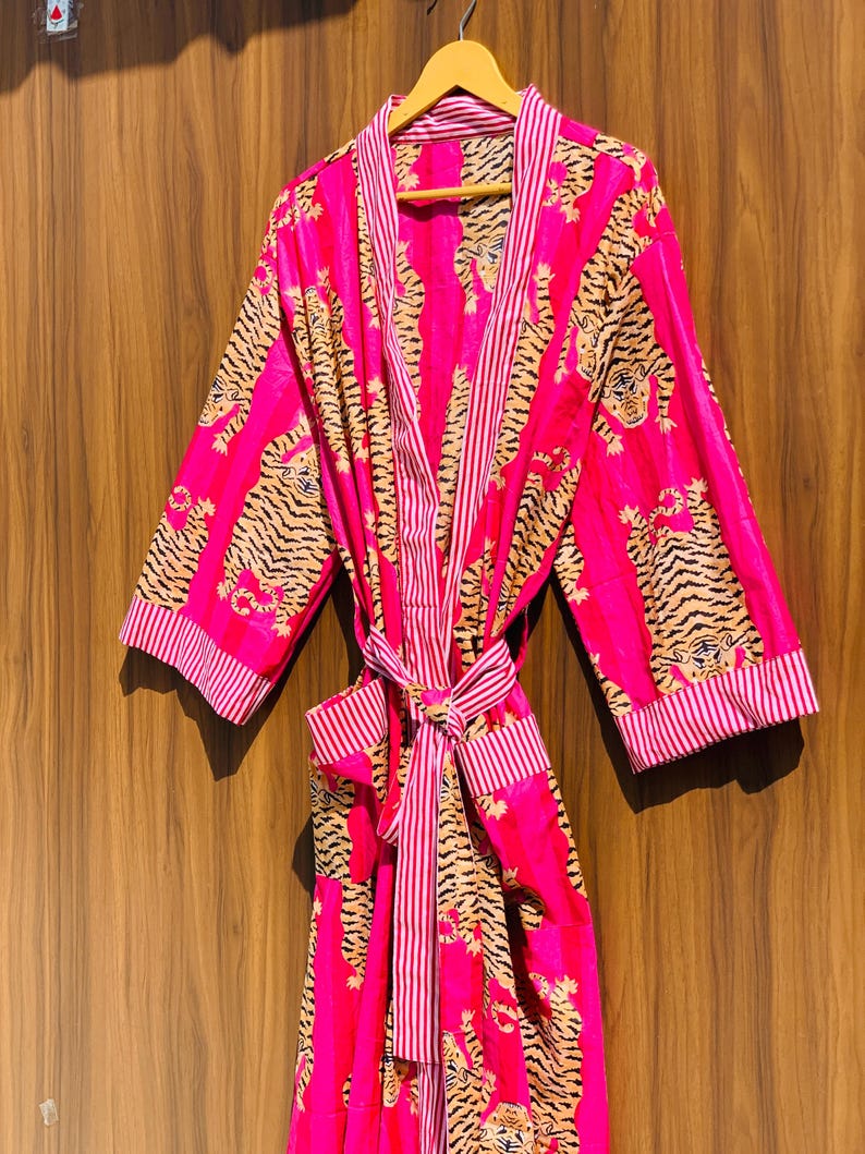 May include: A vibrant pink robe with a tiger print design. The robe features a striped trim and a matching belt. The tiger pattern is in shades of tan and black. The robe is hanging on a wooden hanger.